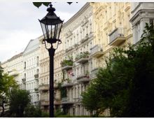 Reihe von historischen, reich verzierten Mietshäusern mit dekorativen Fassaden und Balkonen, flankiert von grünen Bäumen; im Vordergrund eine schwarze Straßenlaterne im traditionellen Stil.