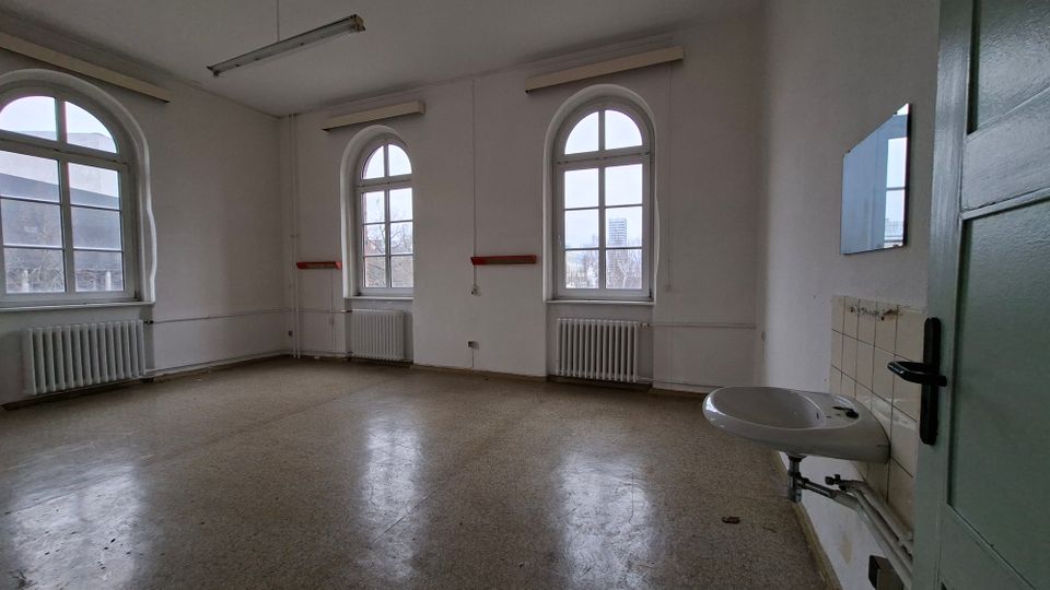 Leerer, schlicht gehaltener Innenraum mit hohen weißen Wänden, drei großen Rundbogenfenstern und Heizkörpern darunter, beleuchtet durch Deckenleuchten. Rechts sind eine Tür und ein wandmontiertes Waschbecken mit Fliesenrückwand zu sehen.