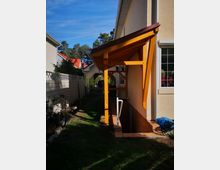 Seitenansicht eines Wohnhauses mit hellem Putz, einem gelb-orangefarbenen Holzvordach und einer Treppe, die in einen Kellerabgang führt. Der Bereich ist von einem Garten mit Rasen, Pflanzen und einem Zaun umgeben, im Hintergrund sind weitere Häuser und Bäume sichtbar.