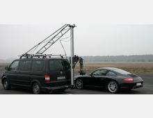 Ein schwarzer Van mit befestigtem Kamerakran steht neben einem schwarzen Sportwagen auf einer asphaltierten Fläche, in einer offenen Landschaft mit Wald im Hintergrund. Der Kamerakran scheint für Filmaufnahmen konzipiert zu sein.