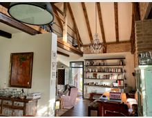 Gemütliches Wohnzimmer mit offenem Dachgebälk, Backsteinwand und einem Kronleuchter. Der Raum verfügt über eine Bücherwand, einen hölzernen Schreibtisch mit Laptop, eine Sitzecke sowie Dekorationen wie Gemälde und Gläser auf einem rustikalen Sideboard.