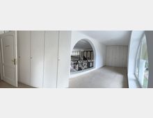 Ein heller Innenraum mit einem offenen Durchgang, der zu einem Bereich mit verzierten Geländern und Säulen führt. Rechts befinden sich große Fenster mit Blick ins Freie, links ein geschlossener Einbauschrank und eine Tür mit der Aufschrift "Direkt.-Raum".