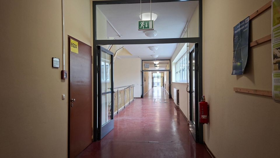 Langer Innenflur eines Schul- oder Bürogebäudes mit roten Linoleumboden, beigen Wänden und Deckenleuchten, durch eine Glastür mit darüberhängendem Notausgangsschild. Rechts verläuft eine Fensterreihe mit Heizkörpern, daneben hängen Aushänge; an der Wand ist ein Feuerlöscher montiert.