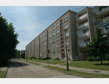 Wohnblock mit mehreren Stockwerken in simpler, rechteckiger Architektur, flankiert von Gehwegen und einer schmalen Straße. Rechts sind einige Bäume und Grünflächen sichtbar, während links der Straßenrand von dichtem Baumbestand gesäumt wird.