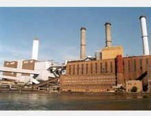 Industrieanlage mit mehreren hohen Schornsteinen, großen Backstein- und Betongebäuden sowie Rohrleitungen, gelegen an einem Flussufer unter klarem Himmel.