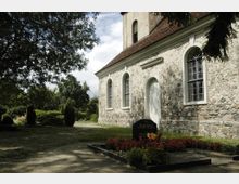 Steinmauer einer historischen Kirche mit Rundbogenfenstern, umgeben von einem Friedhof mit Blumenbeeten und Grabsteinen, eingebettet in eine grüne, baumreiche Landschaft.