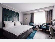 Helles Hotelzimmer mit einem großen Doppelbett, dessen Kopfteil aus dunklen, rustikalen Holztüren besteht. Im Raum befinden sich zwei blaue Sessel, ein Beistelltisch, eine Stehlampe und ein Schreibtisch mit einer Kaffeemaschine; Tageslicht fällt durch ein großes Fenster mit grauen Vorhängen.