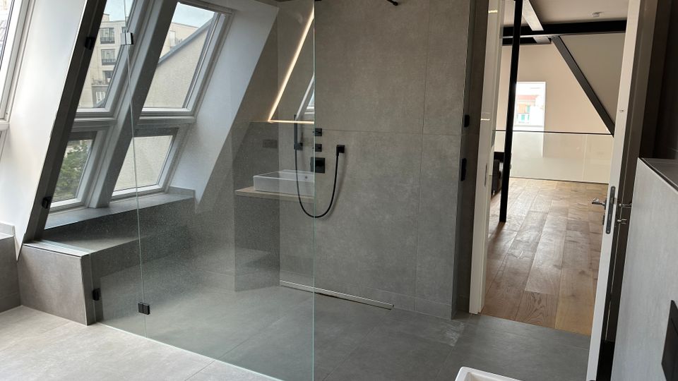 Modernes Badezimmer mit bodengleicher, verglaster Walk-in-Dusche vor einer grauen Fliesenwand und schwarzen Armaturen. Links sorgen große Dachfenster mit Sitzbank für Tageslicht, rechts führt eine offene Tür in einen Flur mit Holzfußboden.