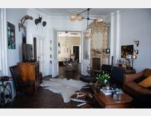 Wohnzimmer mit klassischen und modernen Elementen, darunter ein verzierter Kamin, ein Ledersofa, Holzmöbel und eine Designer-Lampe mit mehreren Glühbirnen. Im Hintergrund ist ein Esszimmer durch doppelflügelige weiße Türen sichtbar, verziert mit hohen Decken und Stuck.