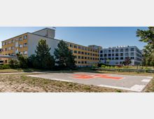 Krankenhausgebäude mit gelbem und weißem Fassadenanstrich, umgeben von Bäumen und Sträuchern. Im Vordergrund befindet sich ein Helikopterlandeplatz mit einem roten „H“-Symbol auf grauem Bodenbelag.