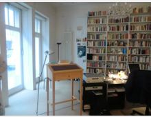 Ein heller Arbeitsraum mit großen Fenstern auf der linken Seite, einer Bücherwand rechts und einem Schreibtisch mit Lampen, Computer und Büromaterial. Im Vordergrund steht ein kleines hölzernes Pult, darüber ein Kronleuchter.