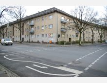 Kreuzung vor einem mehrstöckigen Wohngebäude aus beige-braunem Putz mit Flachdächern und kleinen Balkonen. Entlang der Straße stehen kahle Bäume, und Verkehrsmarkierungen sowie ein Radweg sind sichtbar.