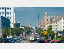Straßenansicht in einer urbanen Umgebung mit dichtem Verkehr, umgeben von Wohn- und Bürogebäuden. Im Hintergrund ist der Berliner Fernsehturm sowie weitere historische Gebäude zu sehen.