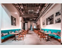 Moderner Innenraum eines Cafés mit hohen Decken und sichtbaren Leitungen. Entlang beider Wände befinden sich türkisfarbene Sitzbänke mit bunten Kissen, ergänzt durch Holztische und orangefarbene Stühle; an den Wänden hängen schwarz-weiße Fotokunstwerke.