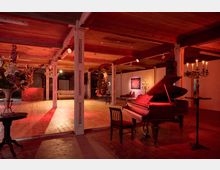 Ein Veranstaltungsraum mit Holzboden und freiliegenden Deckenbalken, beleuchtet durch warmes, rotes Licht. Im Vordergrund steht ein Flügel mit Stuhl, während im Hintergrund dekorative Elemente wie Blumengestecke, Kerzenständer und ein Sofa zu sehen sind.