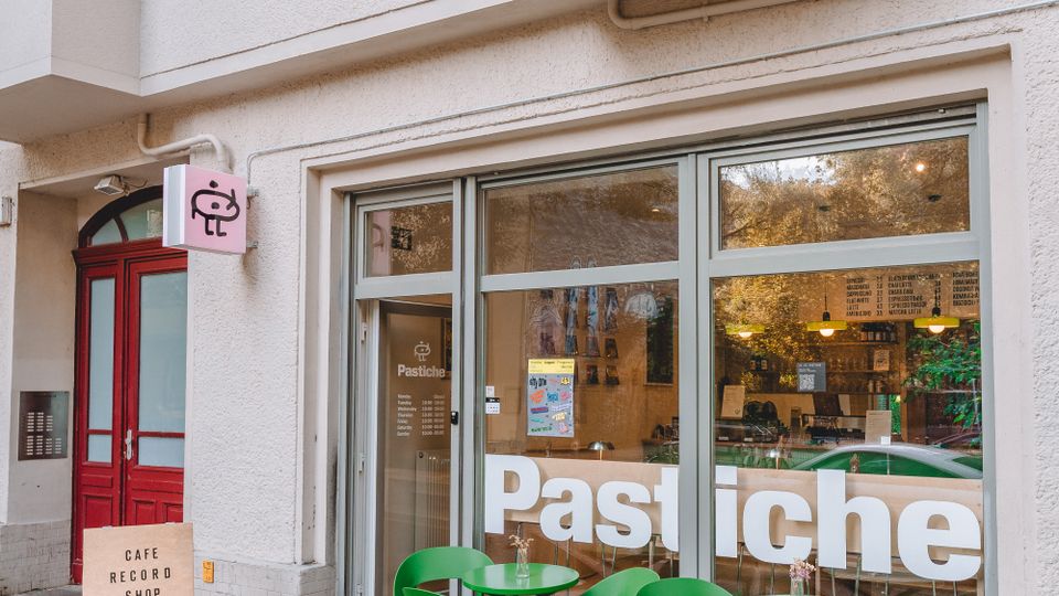 Street-level view of a cafe storefront in a light stucco apartment building, with large front windows labeled “Pastiche” and a small sidewalk seating area of green chairs and round tables. A red entry door and a small hanging sign are beside the windows, with balconies above.