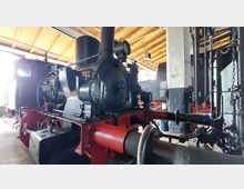 Eine historische Dampflok in einem Lokschuppen mit Holzbalkendecke, weiß gestrichenen Wänden und technischen Geräten im Hintergrund. Die Lokomotive ist schwarz mit roten Elementen und trägt die Aufschrift "Pritzwalk".