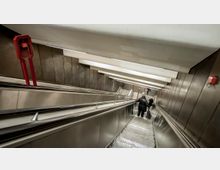 Eine lange, beleuchtete Rolltreppe in einer U-Bahn-Station mit dunklen Wandverkleidungen aus Metall und roten Notrufkästen an den Seiten. Zwei Personen stehen auf der Rolltreppe, eine mit einem Rollkoffer.