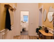 Ein schmaler Flur mit gelben Wänden und Holzfußboden, ausgestattet mit einer Garderobe an der linken Wand und einer Holzkonsole mit Dekorationen und einer Pflanze rechts. Am Ende des Flurs ist ein Badezimmer mit weißer Einrichtung, einem Fenster und gefalteten Handtüchern sichtbar.