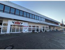 Zweistöckiges Gebäude mit weißer Fassade und großen Fenstern im oberen Bereich, im Erdgeschoss befinden sich ein Kampfsportstudio („Club Sensei Kampfsport“) und ein Autohaus („Hans Laatizg Automobile“). Vor dem Gebäude gibt es einen gepflasterten Vorplatz mit Pflanzkübeln.