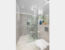 Modernes Badezimmer mit großformatigen grauen Fliesen, bodengleicher Glasdusche mit Handbrause und Ablaufrinne sowie großer Spiegelwand. Rechts sind eine wandhängende Toilette, ein Spülkastenpanel und eine Ablage mit blauen Vasen und Körben zu sehen.