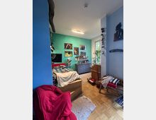 Ein gemütlich eingerichtetes Schlafzimmer mit Parkettboden, einer türkisgrünen Akzentwand, an der gerahmte Bilder hängen, und einer Kommode. Im Vordergrund steht ein Holzbett mit bunten Decken, daneben ein roter Sessel und ein kleiner Teppich.