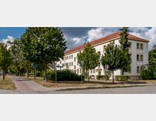 Mehrstöckiges Wohngebäude mit hellbeiger Fassade und roten Dachziegeln, umgeben von Bäumen und Sträuchern. Im Vordergrund verläuft ein gepflasterter Geh- und Radweg entlang einer Straße mit Tempo-30-Schild.