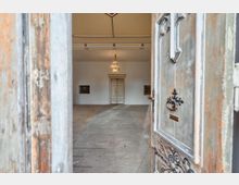 Blick durch eine halb geöffnete, verwitterte Holztür in einen hohen, hellen Innenraum mit Stuckdecke und Kronleuchter. Im Hintergrund steht eine doppelflügelige Tür an der Stirnwand, flankiert von zwei gerahmten Bildern.