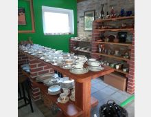 Raum mit Regalen aus Ziegelsteinen und Holz, die verschiedene Geschirrteile wie Tassen, Teller und Gläser präsentieren; im Vordergrund ein Holztresen mit weiterem Porzellan, im Hintergrund eine grüne Wand mit einem kleinen Fenster.