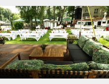 Außenbereich mit Holzterrasse und Lounge-Sofa im Vordergrund, dahinter eine Rasenfläche mit vielen weißen, überdachten Bänken und Tischen in Reihen. Umgeben von hohen Bäumen, Sonnenschirmen und kleinen Gebäuden im Hintergrund.