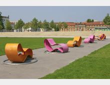 Ein moderner Park mit geschwungenen, bunten Liegebänken in Pink und Orange auf einem gepflasterten Weg, umgeben von Rasenflächen. Im Hintergrund befinden sich Bäume, historische Gebäude mit roten Dächern und eine breite Freitreppe.