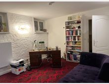 Ein Arbeitszimmer mit weiß gestrichener Backsteinwand, einem klassischen Holzschreibtisch mit einer Schreibtischlampe und einem Computer. Im Hintergrund befinden sich ein weißes Regal mit Büchern und Dekorationsobjekten, offene Türen, eine Heizung sowie zwei kleine Fenster. Ein Teppich mit rotem Muster bedeckt den Boden, und auf der rechten Seite steht ein dunkelviolettes Sofa.