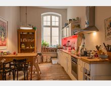 Gemütliche Wohnküche mit Holzdielenboden, einem Holztisch mit Stühlen, offenen Regalen und einer modernen Einbauküche an der rechten Wand. Ein großes Fenster in der Mitte lässt Tageslicht herein, während Pflanzen und Bücher die Fensterbank dekorieren.