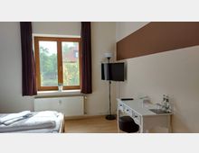 Ein schlichtes Schlafzimmer mit einem großen Holzrahmenfenster, daneben ein Fernseher an der Wand. Ein weißer Schreibtisch mit einem Hocker, einem Fernsehgerät, Gläsern und Wasserflaschen steht an der rechten Wand, während das Fenster einen Blick nach draußen auf ein Haus und Bäume bietet.