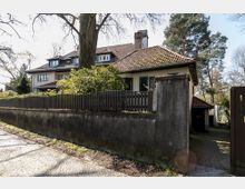 Wohnhaus mit Satteldach und verputzter Fassade, umgeben von einem hohen Gartenzaun und einer gemauerten Mauer; im Vordergrund Bürgersteig aus Kopfsteinpflaster, im Hintergrund Bäume.