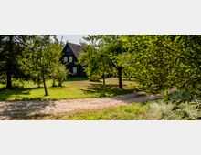 Holzhaus mit Satteldach inmitten eines Gartens mit Bäumen und Rasenflächen, im Vordergrund ein geschwungener, unbefestigter Weg.