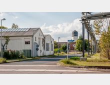 Industriegebiet mit einer Straße, die von einem verfallenen weißen Gebäude und einem hohen Wasserturm im Hintergrund flankiert wird. Rechts verlaufen erhöhte Rohrleitungen parallel zur Straße, und Verkehrsschilder, wie ein "30 km/h"-Schild und ein Bahnübergangsschild, sind sichtbar.