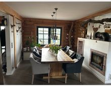 Wohnzimmer in rustikalem Stil mit Holzwänden und Holzmöbeln. Zu sehen sind ein langer Esstisch mit gepolsterten Stühlen, ein Kamin mit Backsteinverkleidung und dekorative Elemente wie hängende Lampen, Pflanzen und Weinflaschenhalter.