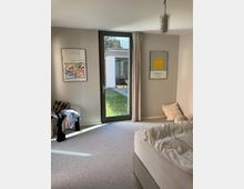 Schlafzimmer mit einem großen Glastürelement, das in einen Garten führt. Der Raum ist mit einem Bett, einem Stuhl mit Kleidung, zwei gerahmten Bildern an der Wand und einem Teppichboden ausgestattet.