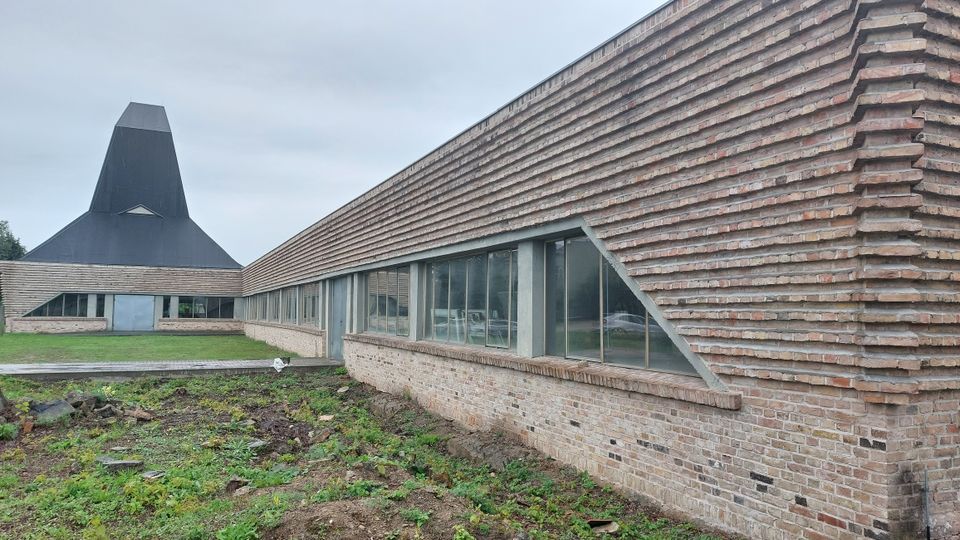 Modernes Gebäude mit einer markanten Ziegelfassade und großen rechteckigen Fenstern; im Hintergrund ein Turm mit einer schwarzen, spitz zulaufenden Dachkonstruktion. Die Umgebung besteht aus einer Grünfläche und einem unbebauten, teils bewachsenen Erdboden.
