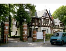 Historisches Gebäude im Fachwerkstil mit weißen Wänden und dunklen Holzelementen, umgeben von Bäumen. Im Vordergrund befinden sich ein gepflasterter Eingang mit gemauerten Torpfeilern und eine Informationstafel.