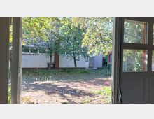 Blick aus einer offenen Tür auf einen gepflasterten Hof mit vereinzeltem Grasbewuchs, mehreren Bäumen und einer grauen Gebäudefassade im Hintergrund. Sonnenlicht fällt durch das Laub und erzeugt helle Schattenmuster.