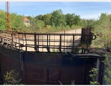 Rostige Stahlkonstruktion eines ehemaligen Industriegebäudes oder Beckens, umgeben von dichter Vegetation und Bäumen; im Hintergrund sind ein Gebäude mit rotem Dach und ein blauer Himmel sichtbar.