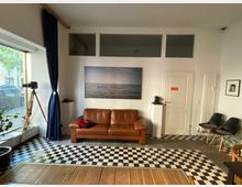 Wohnzimmerartiger Raum mit einer braunen Ledercouch an einer weißen Wand, darauf ein großes Foto des Meeres. Links ein großes Fenster mit dunkelblauen Vorhängen und einem Stativ, rechts eine weiße Tür und schwarze Stühle. Der Boden besteht aus schwarz-weißen Fliesen und Holzelementen.