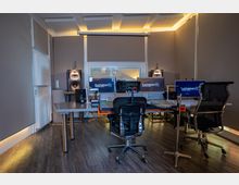 Tonstudio mit einer professionellen Ausstattung: zwei große Studiomonitore, mehrere Bildschirme, ein Mischpult sowie Mikrofone und Lautsprecher. Der Raum hat schallisolierte Wände, einen Holzfußboden und eine zentrale Arbeitsstation mit Bürostühlen.