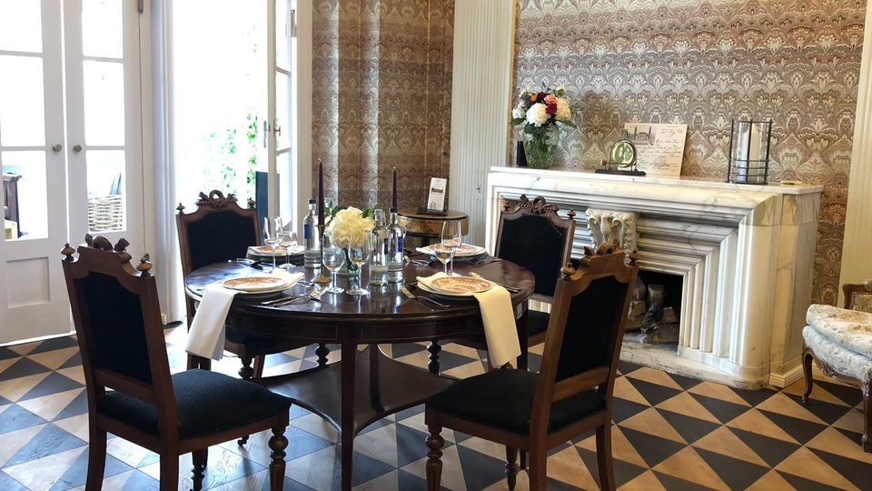 Elegantes Speisezimmer mit rundem Holztisch und vier gepolsterten Stühlen, gedeckt mit Gläsern und Flaschen. Im Hintergrund stehen hohe Fenstertüren, eine gemusterte Tapete und ein weißer Marmorkamin; der Boden ist mit schwarz-beigen Rautenfliesen ausgelegt.