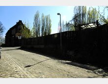 Straßenszene mit Kopfsteinpflaster, links ein altes Backsteingebäude mit spitzem Dach, daneben eine Holzzaunreihe und hohe, schlanke Bäume. Im Vordergrund steht eine Straßenlaterne, im Schattenbereich sind Teile eines Fahrrads sichtbar.