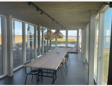Moderner Innenraum mit großem Holztisch und Stühlen, umgeben von bodentiefen Fenstern mit Blick auf eine Landschaft aus Schilf und Wasser. Die Decke ist aus Beton, und mehrere geflochtene Lampen hängen über dem Tisch.