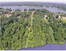 Luftbild einer von Wald bedeckten Halbinsel mit einem schmalen Weg, der zu einem Steg am Wasser führt, umgeben von einem großen See. Im Hintergrund sind ein Dorf mit Häusern und mehr Bäume sichtbar.