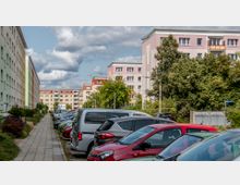 Reihen von farbigen Plattenbauten mit vier bis fünf Stockwerken, flankiert von einem Gehweg und einer Parkfläche, die mit Autos gefüllt ist; rechts befinden sich Bäume und Müllcontainer.